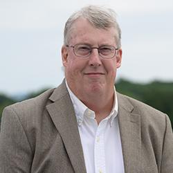 caucasian man, gray hair, glasses, wearing white button up shirt and brown suit jacket